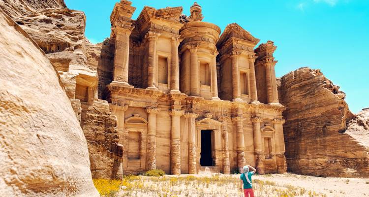 Un visiteur se tenant devant la majestueuse facade en grès sculptée du Monastère à Petra sous un ciel bleu éclatant