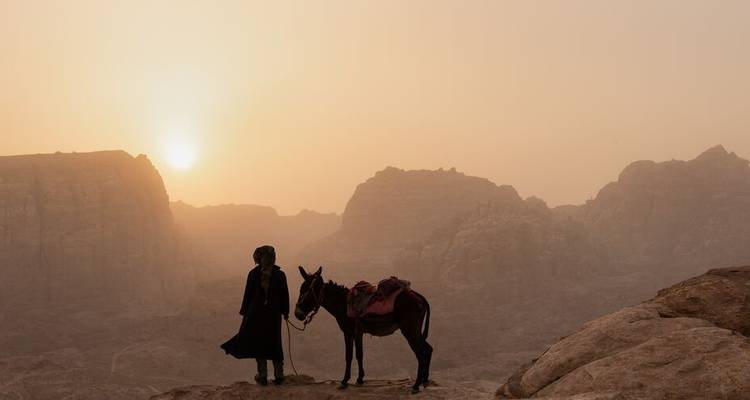 Silhouette d'une personne et d'un âne sur une crête rocheuse au coucher de soleil poussiéreux au-dessus de montagnes désertiques accidentées