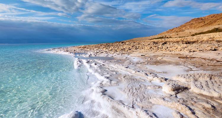 Kristalhelder turquoise water klapt tegen witte zoutformaties langs de Dode Zee-kust onder een uitgestrekte hemel.