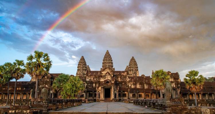 Les tours emblématiques d'Angkor Wat sous un ciel dramatique couronné d'un arc-en-ciel éclatant et encadré par des palmiers