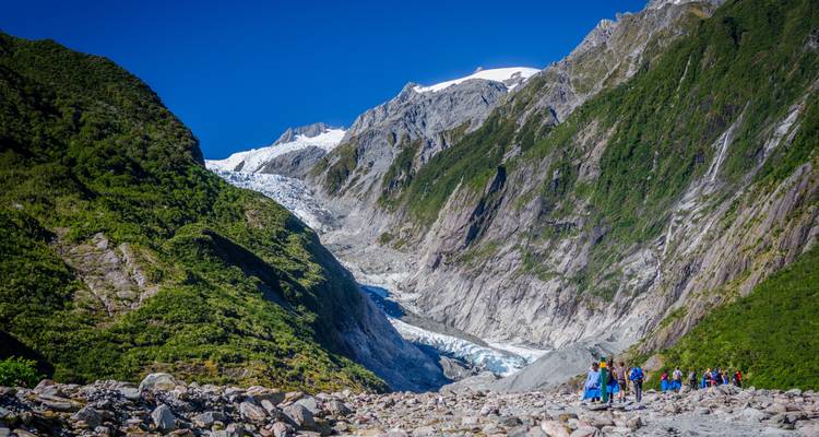 Un grupo guiado vistiendo equipo de seguridad se encuentra de pie en la morrena rocosa frente al imponente Glaciar Franz Josef bajo un cielo azul despejado.