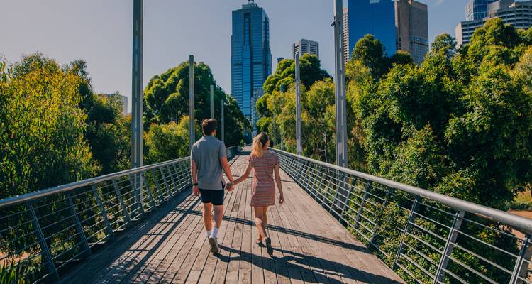 Un jeune couple se tenant par la main traverse un pont en bois en direction de l'horizon urbain moderne de Melbourne encadré par un parc verdoyant.