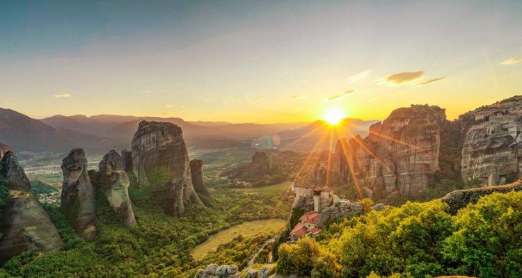 Zonsopgang boven imposante rotspilaars en kloosters van Meteora met een weelderige vallei daaronder.