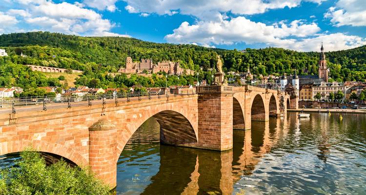 El histórico Puente Viejo se arquea sobre el río Neckar con el castillo de Heidelberg y una colina boscosa detrás.