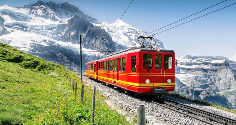 Tren de engranaje rojo brillante subiendo por una vía de montaña con picos alpinos cubiertos de nieve de fondo bajo un cielo azul despejado.