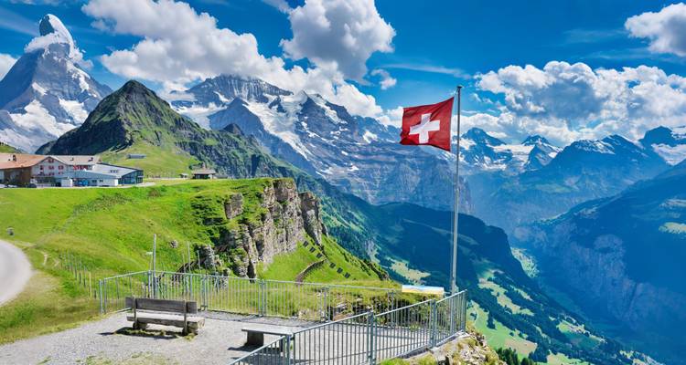 Vista alpina panorámica con la bandera suiza ondeando en una plataforma de mirador que da a dramáticas montañas nevadas y un verde valle.