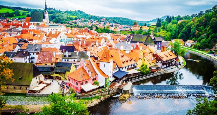 Panorama aéreo colorido del pueblo medieval de Cesky Krumlov con tejados rojos, rodeado por un río sinuoso bajo cielos nublados.