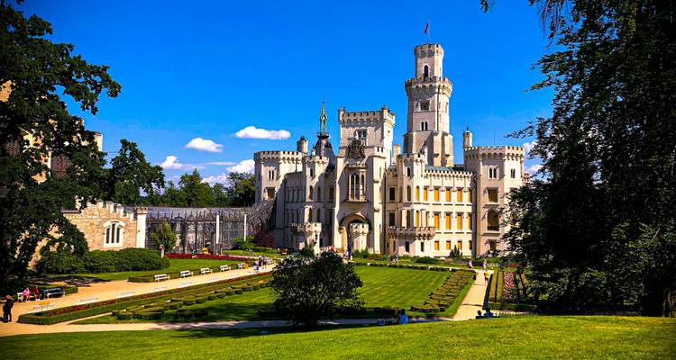 El majestuoso Castillo Hluboka de estilo Neo-Gótico enmarcado por jardines bien cuidados contra un cielo azul vibrante.