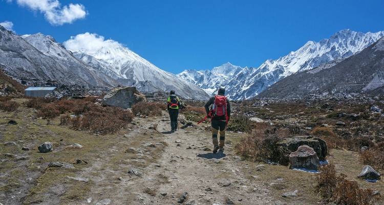 Dos trekkers con mochilas avanzan por un sendero rocoso del Himalaya hacia imponentes picos nevados bajo un cielo azul profundo.