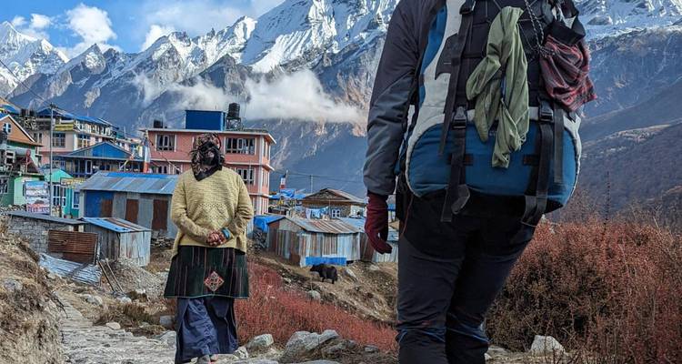 Mochilero con equipo pasa junto a una mujer local abrigada en una calle de piedra con casas de montaña en colores pastel y picos nevados a lo lejos.