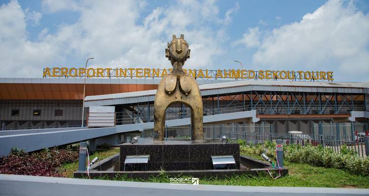 Entrada principal del aeropuerto de Conakry con fachada de cristal moderno y una gran estatua dorada.