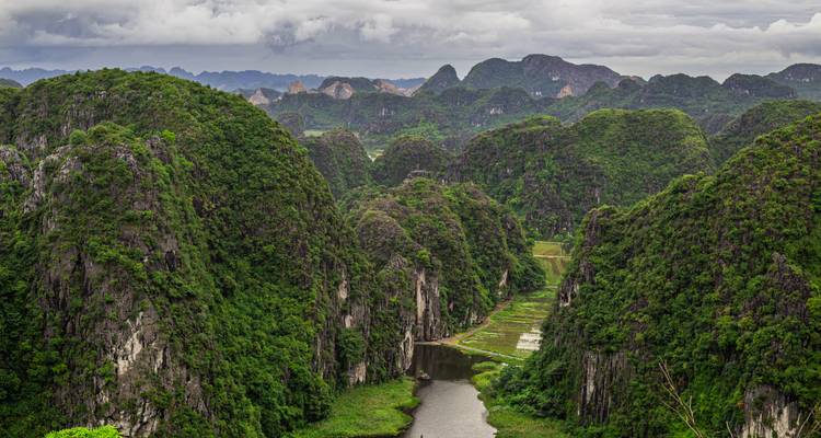 Des formations calcaires karstiques imposantes, recouvertes de jungle, s'élèvent d'une vallée fluviale sinueuse à Ninh Binh sous des nuages gris menaçants.
