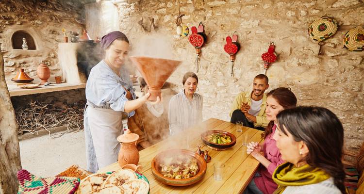 Ein lächelnder Gastgeber präsentiert ein dampfendes Tajine für Gäste, die an einem rustikalen Tisch mit Steinmauern sitzen und die marokkanische Küche geniessen.