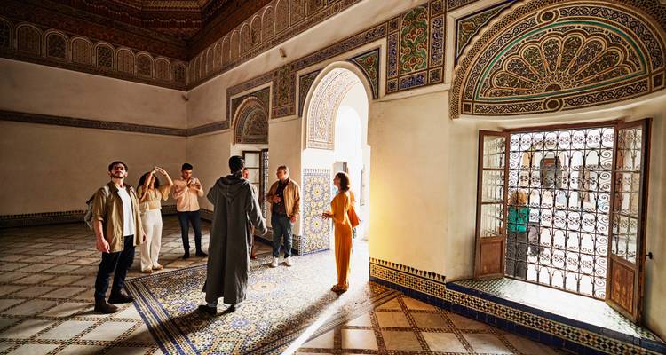 Besucher erkunden eine aufwendig geflieste Halle eines Palastes in Marrakesch und bewundern kunstvolle Bögen und Mosaiken.
