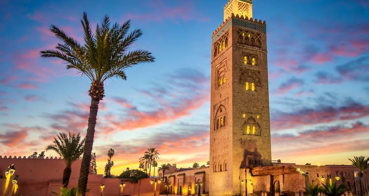 Das Minarett der Koutoubia-Moschee erhebt sich gegen einen lebhaften Himmel bei Sonnenuntergang mit Palmen und leuchtenden Lampen im Vordergrund.