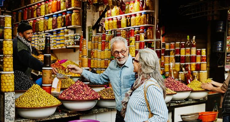 Ein älteres Ehepaar kauft bunte Oliven in einem geschäftigen Marrakesch-Souk, der mit hohen Gläsern voller eingelegter Gemüse gesäumt ist.