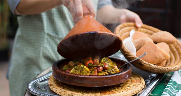 Nahaufnahme eines dampfenden marokkanischen Tajine, das neben frischem Brot auf einer geflochtenen Matte serviert wird.