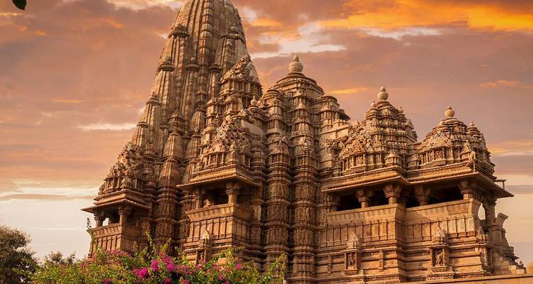 Der verzierte Khajuraho-Tempelturm erstrahlt unter einem leuchtend orangefarbenen Himmel bei Sonnenuntergang, umrahmt von blühender Bougainvillea.