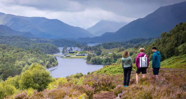 Tres excursionistas de pie en una ladera cubierta de brezo con vistas a un tranquilo lago y capas de montañas azul verdosas en las Tierras Altas de Escocia.
