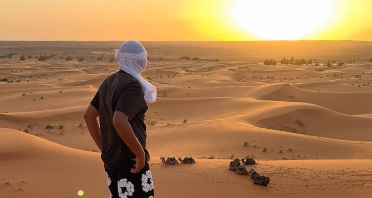 Reiziger met hoofddoek die de zon ziet ondergaan over uitgestrekte woestijnduinen met rustende kamelen eronder.
