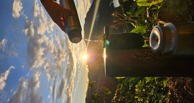 Una mano vertiendo agua caliente de un termo en un filtro de metal con el amanecer sobre montañas verdes de fondo.
