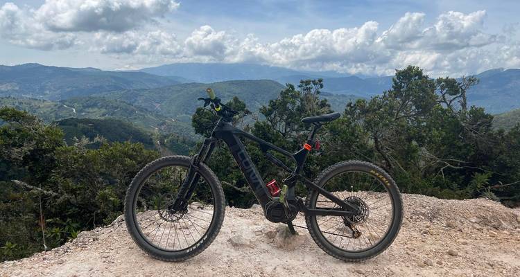 Bicicleta eléctrica de montaña estacionada en un mirador rocoso con vistas panorámicas de las montañas y nubes a lo lejos.