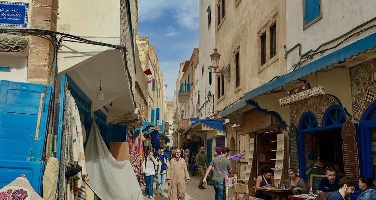 Calle estrecha y concurrida bordeada de tiendas con bordes azules y peatones en una medina marroquí.
