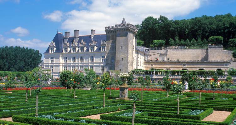 Castillo renacentista formal rodeado de impecables jardines parterre con setos, flores y estatuas bajo un cielo azul.