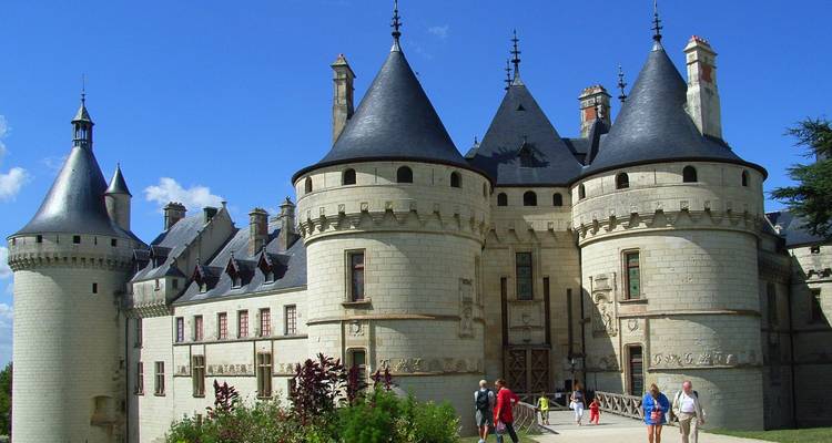 Castillo de cuento de hadas con tejados de pizarra cónica y torres redondas, visitantes paseando cerca de la entrada del puente levadizo.