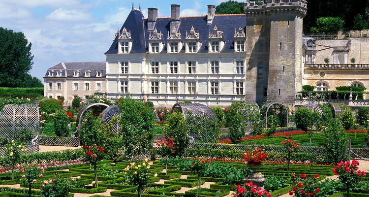 Jardín formal de rosas y setos de boj frente a una larga ala del castillo renacentista bajo un cielo de verano.
