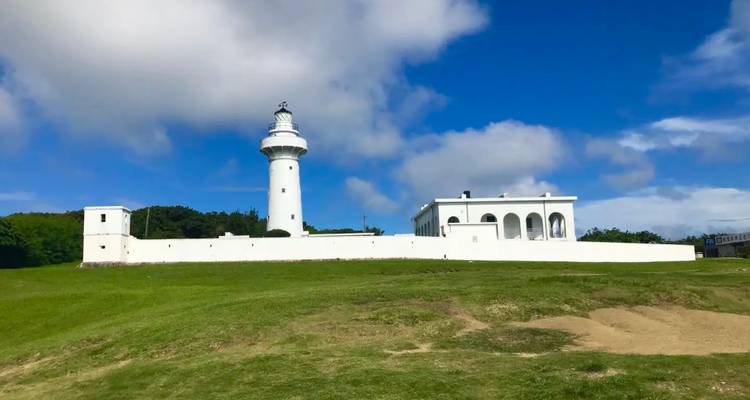 Ein weißer Küstenleuchtturm und ein angrenzendes Gebäude stehen unter einem strahlend blauen Himmel auf einem grasigen Hügel.