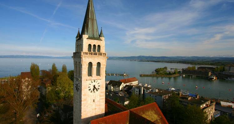 Hoher Kirchturm mit Uhrengesichtern überblickt den Bodensee und die fernen Alpen.
