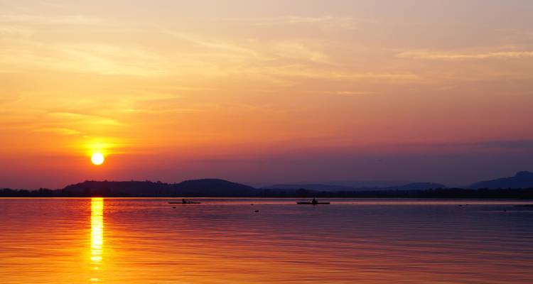Heiterer Sonnenuntergang über ruhigen Seewassern mit orangefarbenen Spiegelungen und entfernten Kajakfahrern.