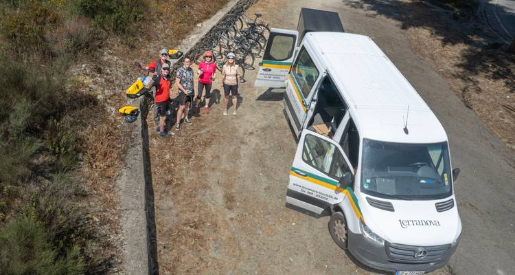 Un grupo de ciclismo está junto a una furgoneta de apoyo y un remolque cargado de bicicletas en un camino rural.