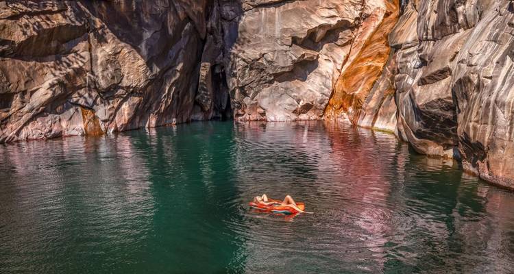 Un viajero flotando tranquilamente boca arriba en una piscina natural de roca verde esmeralda rodeada de paredes de cañón que se elevan hacia el cielo.