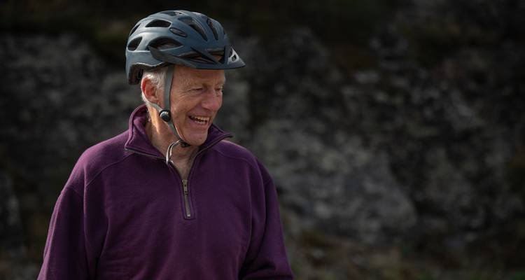 Ein lächelnder älterer Radfahrer in einem Helm und einer violetten Jacke, fotografiert vor einem felsigen Hintergrund.