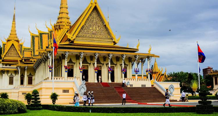 Der prunkvolle Königspalast in Phnom Penh mit goldenen Dächern und kambodschanischen Flaggen, besucht von uniformierten Wachen und Touristen.