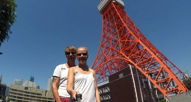 Ein Paar, das ein Selfie mit dem leuchtend roten Tokioer Fernsehturm macht, der sich in einen klaren blauen Himmel erhebt.