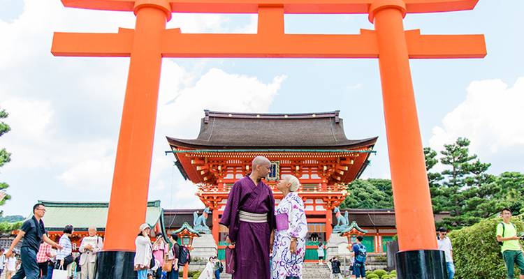 Lebendiges orangefarbenes Torii-Tor mit ältereم Ehepaar in traditioneller Kleidung und Schreingebäuden im Hintergrund.