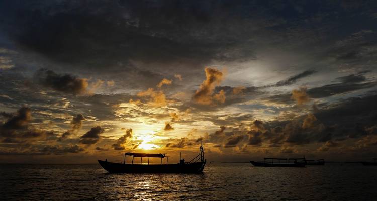 Silhouettierte Holzboote ruhen auf ruhigen Meereswassern unter einem dramatischen orangefarbenen Himmel bei Sonnenuntergang.