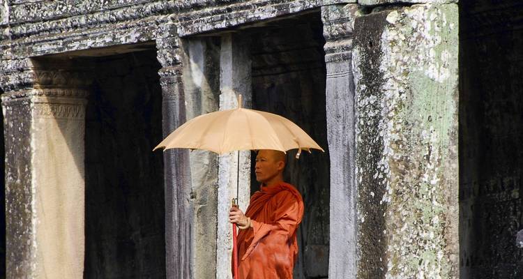 Ein Mönch mit einem Regenschirm steht neben den alten Steinpfeilern eines Angkor-Tempels.