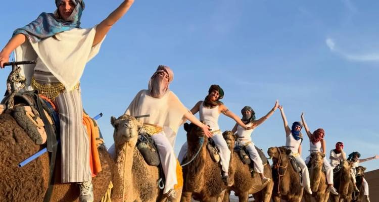 Una fila de mujeres con ropa del desierto cabalgan camellos por dunas doradas, levantando los brazos para celebrar.