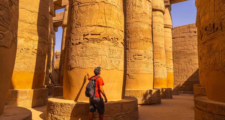 Un voyageur examine d'énormes colonnes couvertes de hiéroglyphes à l'intérieur du temple de Karnak, baigné par la lumière du soleil chaud.