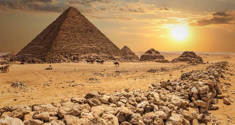 Une pyramide s'élève au-dessus du désert au coucher du soleil avec des chameaux et des cavaliers en silhouette contre le ciel luminescent.