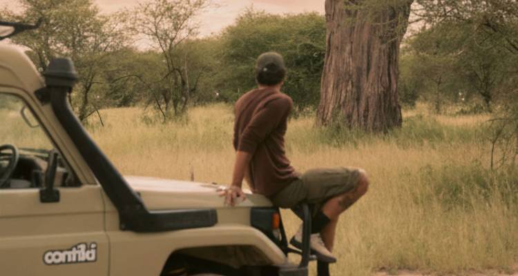 Voyageur assis sur le capot d'un véhicule de safari observant la savane africaine et un arbre ressemblant à un baobab.