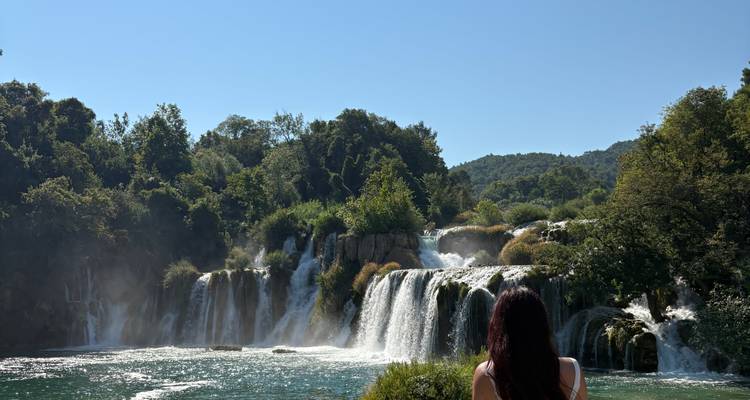 Ein Besucher mit langem dunklem Haar bewundert die Wasserfälle von Krka unter einem klaren blauen Sommerhimmel