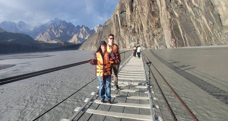 Deux voyageurs portant des gilets de sauvetage se tiennent sur un pont suspendu en bois étroit au-dessus d'une large rivière grise avec des pics déchiquetés en arrière-plan.