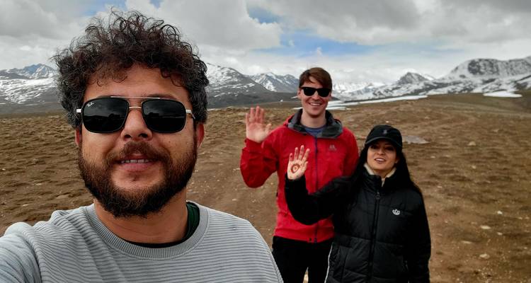 Autoportrait de trois amis agitant la main sur un plateau désertique avec des montagnes enneigées sous un ciel nuageux.