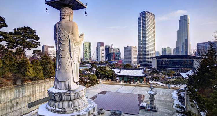Een groot stenen Boeddhabeeld kijkt uit over een met sneeuw bestoven tempelplein met moderne Seoulse wolkenkrabbers erachter.
