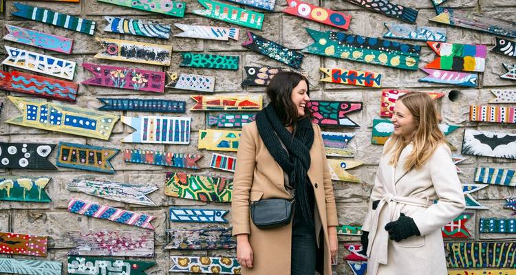 Deux femmes discutent et sourient devant un mur recouvert de pièces d'art en bois peint aux couleurs vives.
